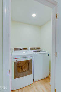 A small laundry room with a white washer and dryer set. The appliances are side by side, under a bright overhead light on a wooden floor. The walls are painted a light color, creating a bright and clean atmosphere.