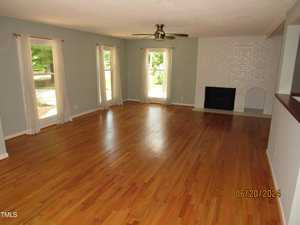 A spacious living room with wooden floors, light blue walls, and white curtains. Two large windows let in natural light. There's a ceiling fan and a white brick fireplace on the right wall.