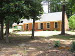 A charming brick house at 111 Ridge Road in Butner boasts a gable roof, surrounded by lush trees and grass. The sun plays across several windows and a covered carport to the left, casting gentle shadows on the lawn. A date stamp reads "06/20/2024.