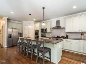 A modern kitchen features white cabinets, a granite countertop, and stainless steel appliances. Three wooden chairs are lined up at the island, and a stainless steel range hood is above the stove. The floor is wooden, adding warmth to the space.
