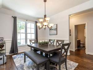 A dining room with a dark wooden table and matching benches. A chandelier hangs above, and a large window with gray curtains lets in natural light. A painting is on the wall, and a hallway leads to a room with a visible sink and toilet.