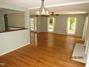 A spacious, open-concept living room with light wood flooring and soft green walls. It features a ceiling fan, large windows with white curtains providing natural light, and a visible open doorway leading to another room. A chandelier hangs from the ceiling.