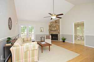 Spacious living room with high ceiling, featuring a striped sofa, coffee table on a rug, and a stone fireplace. Ceiling fan overhead and French doors leading outside. A separate dining area is visible through an open doorway. Natural light brightens the space.