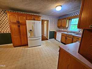 A kitchen with wooden cabinets and patterned wallpaper. There's a white refrigerator, a dishwasher, and a counter with a window above it. The floor is covered with light-colored linoleum, and a ceiling light illuminates the space.