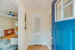 A cozy home entrance with a blue front door. The hallway leads to a living room with a ceiling fan and a glimpse of a kitchen in the background. A small white closet is present, and a partial view of a colorful rug and sofa is visible.