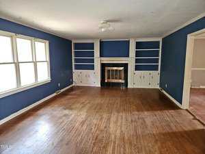 A spacious living room with wooden floors, blue walls, and a white ceiling. There is a ceiling fan and a fireplace with built-in shelving on both sides. Large windows let in natural light. An open doorway leads to another room.