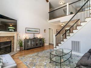 A cozy living room featuring a stone fireplace, a large wall-mounted TV, and a patterned area rug. There's a modern glass coffee table, a decorative cabinet with plants, and a staircase leading to an open upstairs area with black railings.