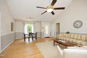 A spacious living room with a high ceiling, featuring a ceiling fan, a striped sofa, a white armchair, and a coffee table on a beige rug. A dining table is near the window. The walls are light-colored, and the flooring is wooden.