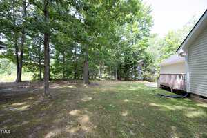 A backyard featuring a grassy area with several large trees providing shade. To the right is part of a light-colored house with a small deck. The scene is tranquil, surrounded by lush greenery and partially sunny skies.
