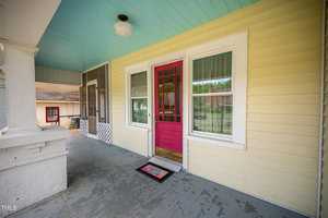 A welcoming wooden porch features a vibrant red door set in a pale yellow house. The porch has a light blue ceiling and white decorative elements, with a doormat at the entrance. Large windows flank the door, offering a glimpse into the interior.