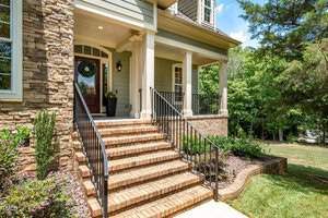 A brick staircase leads up to a house entrance with a covered porch and white columns. The house features a stone and light green exterior. A wreath hangs on the front door, and there are plants and trees in the surrounding yard.