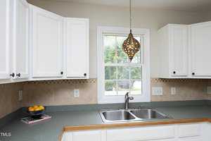 A kitchen with white cabinets, a double sink, and a green countertop. A window above the sink allows natural light in. A decorative hanging light fixture is above the sink, and a bowl of lemons is on the counter.