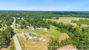 Aerial view of a rural landscape showing a cluster of houses and farm buildings surrounded by greenery and fields. A road runs parallel to the properties, with forested areas and a small pond visible in the distance under a clear blue sky.