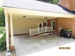 A covered carport with a sloped ramp leading to a side entrance of a brick house. There are three trash bins nearby and it is surrounded by trees. The carport has a shingled roof and two supporting columns. Date stamp in the corner reads 06/20/2024.