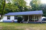 A single-story house at 1618 W B Street, Butner, features light-colored siding and a dark gray roof, nestled amid lush greenery. The cozy porch with chairs and an American flag graces the yard, while a detached garage stands proudly in the background.