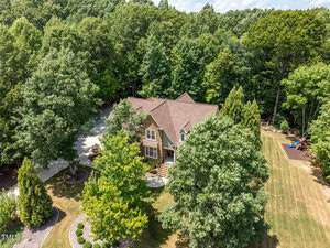 Aerial view of a large house surrounded by dense green trees and a well-maintained lawn. The property includes a driveway and a children's play area at the side. The house has a brown roof and multiple windows.