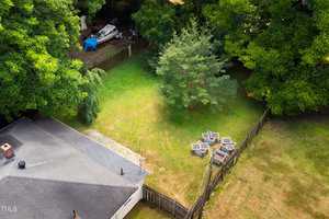 Aerial view of a backyard featuring a grassy area surrounded by trees. A house roof is visible in the lower left corner. In the yard, there's a cluster of chairs arranged around a fire pit and a boat covered with a blue tarp near a fence.