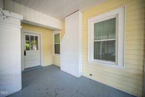 A covered porch with pale yellow siding and a light blue floor. There are two white-framed windows with blinds and a white door with glass panels. A white brick column is in the center of the wall. The ceiling is white paneled.