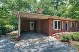 A brick house with a covered porch, featuring a white door and a blue door. It is surrounded by trees and shrubs, with a gravel driveway leading to the porch. A bright, sunny day highlights the greenery.