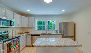 A modern kitchen with white cabinets, granite countertops, and stainless steel appliances. A large window over the sink offers a view of greenery outside. A colorful towel hangs on the oven handle, and a kitchen island stands in the center.