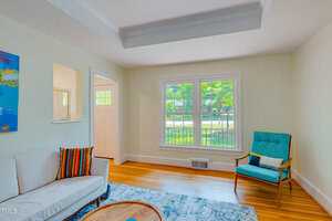 Bright living room with a beige sofa, colorful cushions, and a turquoise armchair. Large window lets in natural light. Hardwood floor with a blue rug. White walls and a small wall art piece add a modern touch.