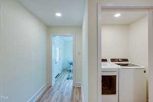 A hallway with light-colored walls and wood flooring leads to a laundry area with a washer and dryer on the right. Natural light enters from a door at the end of the hallway, and a small table is visible in the adjoining room.