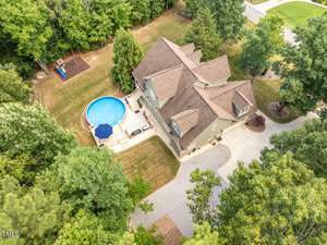 An aerial view of a large house surrounded by trees. The property features a round above-ground pool with a patio and seating area. The driveway leads to the house, and there's a children's playset in the yard.
