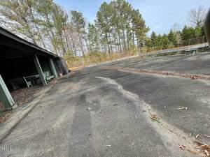 Abandoned go-kart track with cracked asphalt and scattered leaves. Surrounded by tall trees and greenery. A shaded seating area on the left side under a small shelter. The sky is clear and the scene is quiet and deserted.