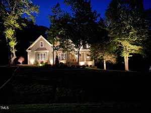 A well-lit house with large windows is surrounded by tall trees at night. The warm lights from the house create a cozy contrast against the dark blue evening sky.