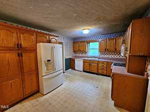 A kitchen with wooden cabinets, a white refrigerator, a dishwasher, an oven, and a microwave. The walls have patterned wallpaper, and a window provides natural light. The ceiling light is on, illuminating the space.