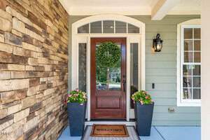 Front porch with a wooden door adorned with a green wreath. Flanked by two large planters with pink flowers. Stone and green siding, black lantern light on the wall. A "Welcome" mat rests on the doorstep.