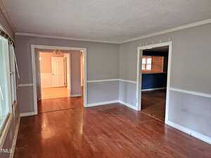 An empty room with light gray walls and wooden floors. A large window is on the left, with adjoining doorways leading to other rooms. One room is brightly lit and another is darker.