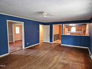 A spacious empty living room with blue walls and hardwood floors. A ceiling fan is centered. The room opens to a kitchen with light wood cabinets and patterned wallpaper. There are two doorways leading to other parts of the house.