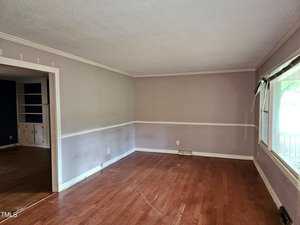 An empty room with light brown hardwood floors, pale walls, and a large window on the right. There's a doorway leading to another room with built-in shelves. The room has a simple white chair rail and a ceiling with crown molding.