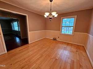A cozy dining room with light wood flooring, beige walls, and white wainscoting. The room features a window on the right and a central chandelier. An open doorway leads to another room with darker wood flooring.