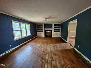 A spacious living room with dark blue walls, hardwood floors, and a white ceiling. There is a large window on the left, built-in shelving on the back wall surrounding a fireplace, and an adjoining room visible on the right.