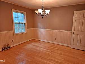 A small, empty room with light wood flooring and beige walls featuring a chair rail. A window with a view of greenery is on the left wall. A white door is partially open on the right, and a ceiling light fixture hangs in the center.