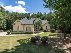 A two-story house at 525 Belmont Circle, with a stone and beige facade, is surrounded by trees in Wake Forest. The manicured lawn features bushes and a circular driveway. A small garden with a rock and plaque rests in the foreground under the clear blue sky.