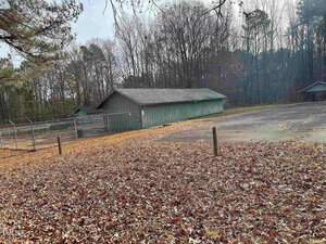 A weathered green building stands near a fenced area and an empty paved lot, surrounded by leaf-strewn ground and bare trees in a wooded area. The sky is overcast, giving the scene a muted, autumnal atmosphere.
