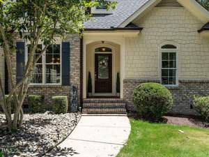 A charming house with a brick and shingle exterior, featuring a central dark wooden door and two tall windows. A path curves through a neatly maintained lawn, accentuated by small shrubs and a tree.