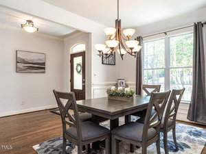 A dining room with a dark wooden table and six chairs. A chandelier with spherical lights hangs above the table. A large window with gray curtains brings in natural light. A framed picture and decorative plant adorn the table. A door leads outside.