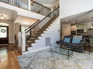A modern living room with a gray sofa, blue and gray pillows, and a glass coffee table on a patterned rug. A staircase with black railings leads to an upper floor. The room is bright, with large windows and a ceiling fan above.