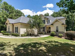 A charming suburban house with light beige siding, dark grey roofing, and brick accents. The property features a well-maintained lawn, small shrubs, and trees, set against a backdrop of tall, lush greenery under a blue sky with scattered clouds.