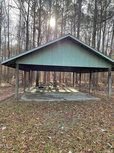 A wooden picnic shelter with a metal roof sits in a forested area. Sunlight filters through the trees, casting shadows on the ground. The shelter contains a picnic table and is surrounded by fallen leaves.
