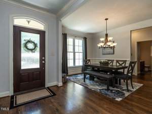A dining room with a dark wooden table, six chairs, and a bench. A chandelier hangs above the table. Large windows with curtains provide natural light. A wreath decorates the front door nearby. Hardwood floors and a decorative rug complete the space.
