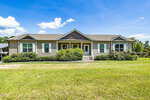 A single-story house at 3218 Old Nc 75 features a gray exterior and green shutters, surrounded by neatly trimmed bushes and a grassy lawn. Under the clear blue sky with a few clouds, steps lead up to the front door, perfectly centered on the facade.