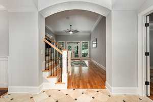 A hallway with a tiled floor leads to a room with wooden flooring. An archway frames a staircase on the left with wooden rails. Beyond, a ceiling fan hangs from the ceiling, and large windows show greenery outside. Doors are on the right.