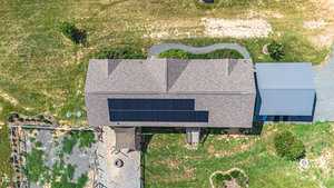 Aerial view of a house with solar panels on the roof. The yard features a defined pathway, a shaded seating area, and a small garden. The surrounding area is grassy with patches of dry ground.