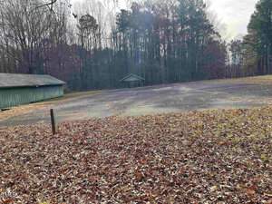 A deserted, leaf-covered parking lot surrounded by tall trees in autumn. Two wooden structures are visible at the edges of the lot, with sunlight filtering through the tree canopy.