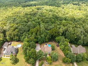 Aerial view of several houses with pools, bordered by a dense forest. The properties are surrounded by green lawns and trees, creating a serene residential area next to the expansive wooded landscape.